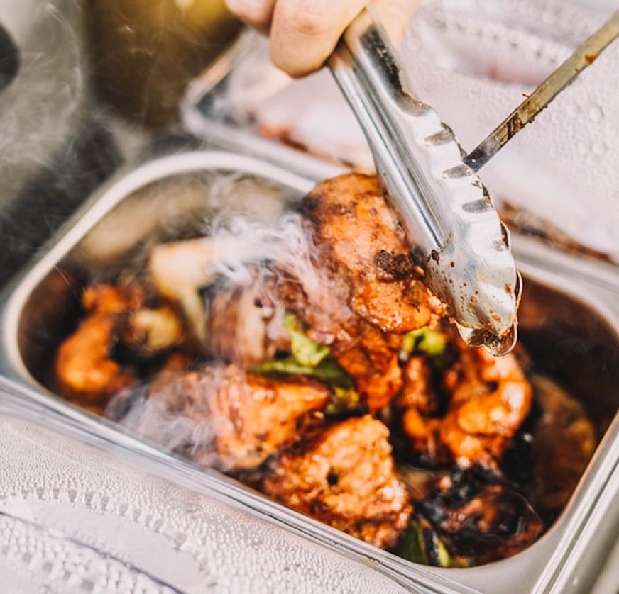 Tongs lift a piece of char-grilled chicken from a steaming stainless catering tray, with fresh basil tucked beneath