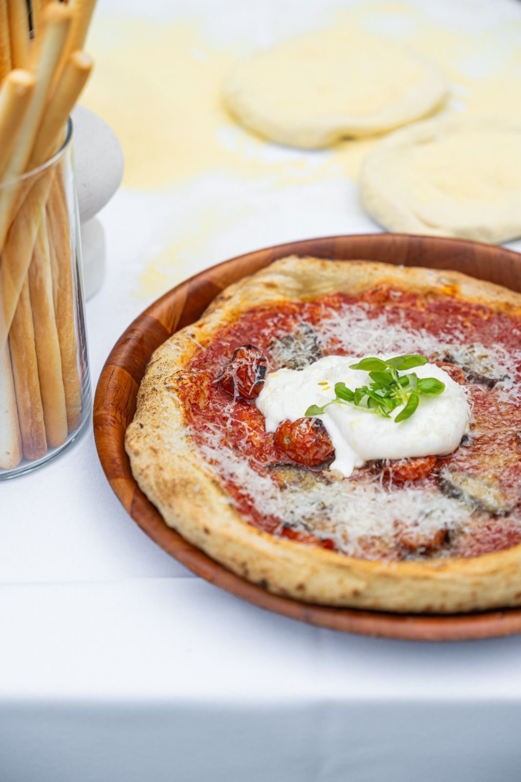 A wood-fired pizza topped with tomato, burrata and basil, served on a wooden board with breadsticks