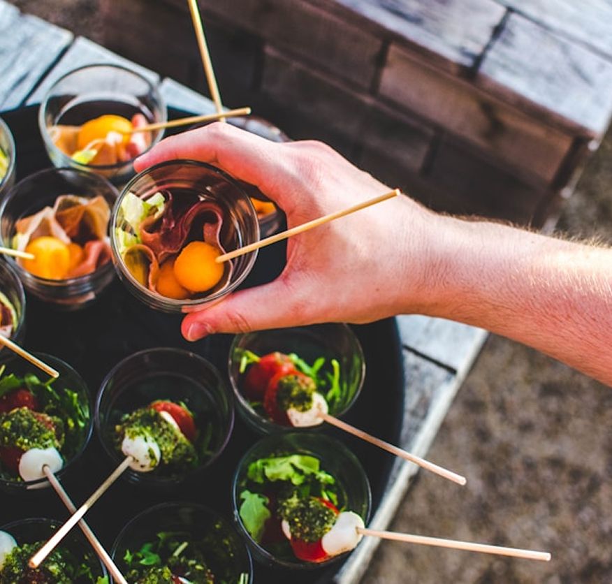 A hand picking up a small glass cup of prosciutto and melon canapé from a tray of individual glass cup canapés, some with tomato, mozzarella and basil skewers, on an outdoor wooden table