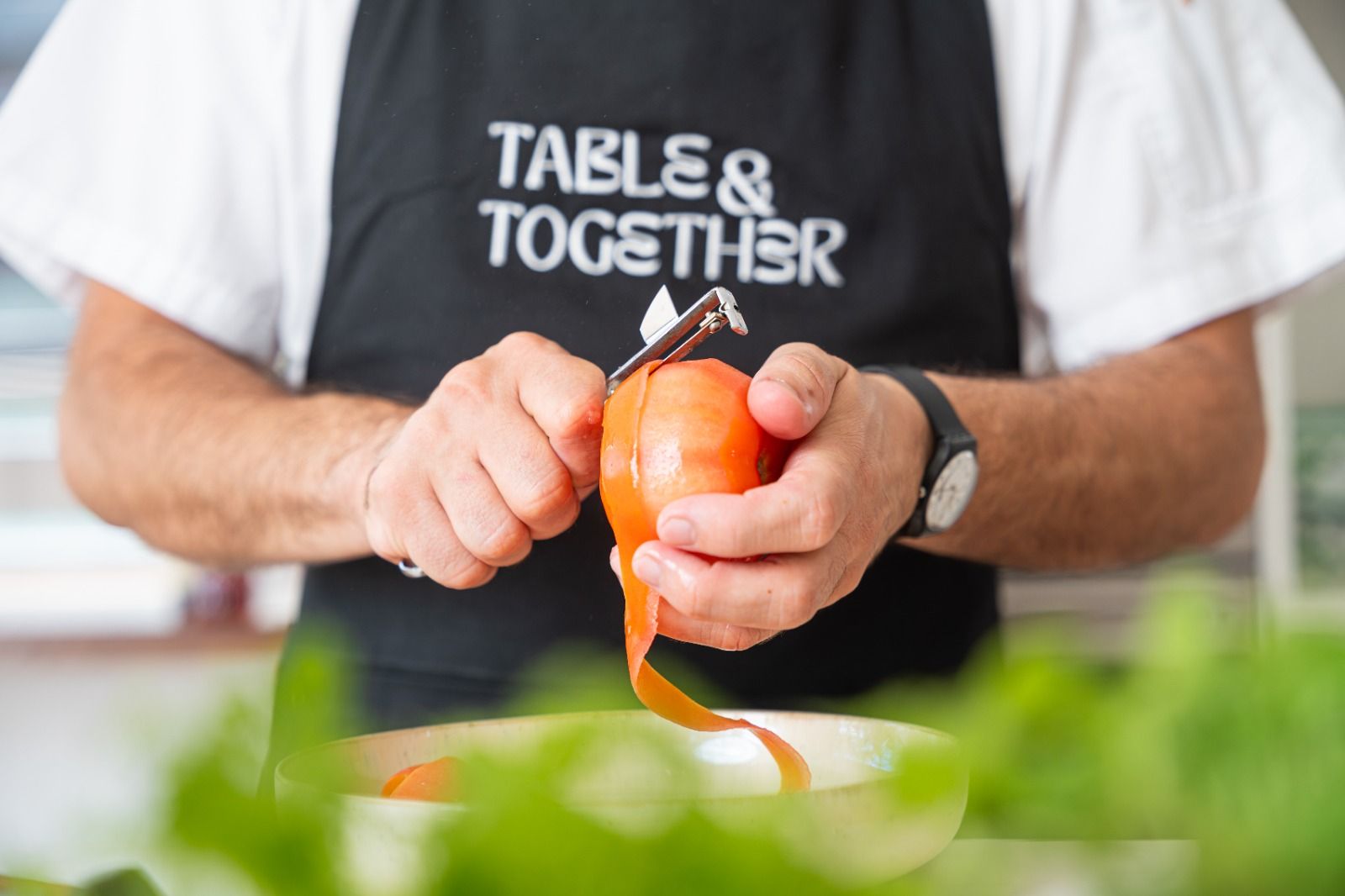 Close-up of hands in a black Table & Together branded apron peeling a tomato with a swivel peeler over a glass bowl, with fresh herbs in the foreground