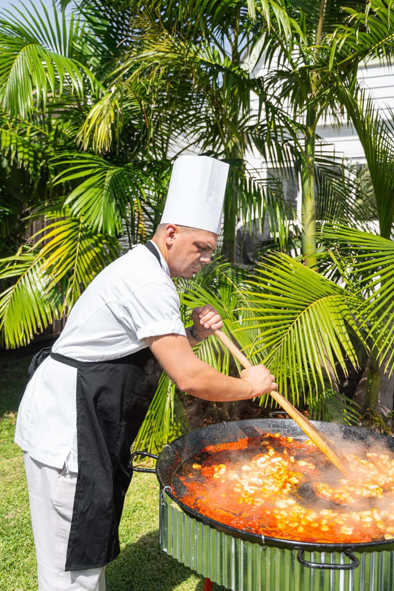A chef in a tall white toque stirs a huge pan of tomato sauce and gnocchi at an outdoor cooking station, palm trees behind him
