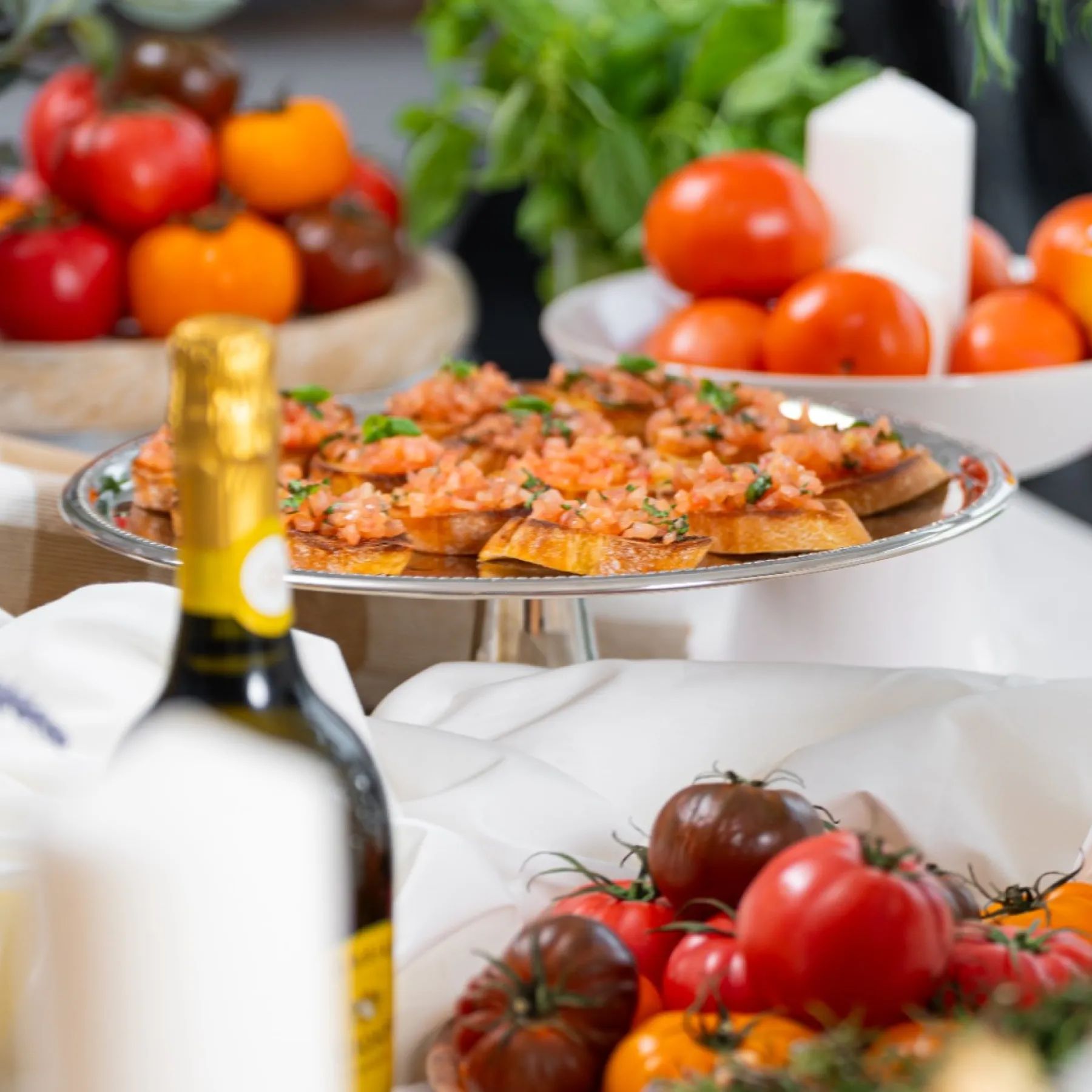 Tomato and basil bruschetta on a silver tray beside heirloom tomatoes, white pillar candles and a bottle of wine, styled for a catered reception