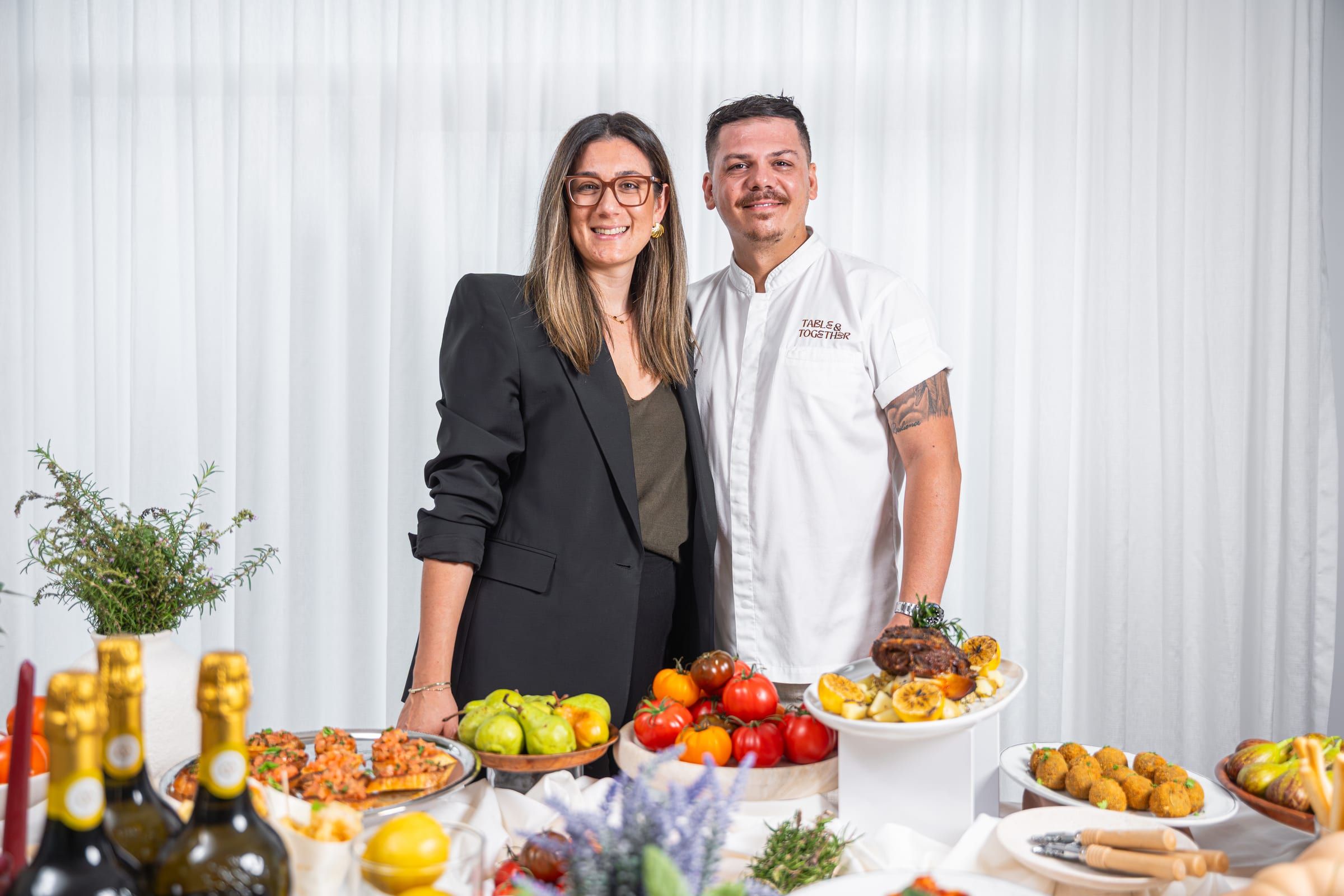 Natasha and Gabriele, the founders of Table & Together, standing behind a table spread with bruschetta, heirloom tomatoes, pears, arancini, a roast joint and prosecco, Gabriele wearing a Table & Together branded chef's shirt