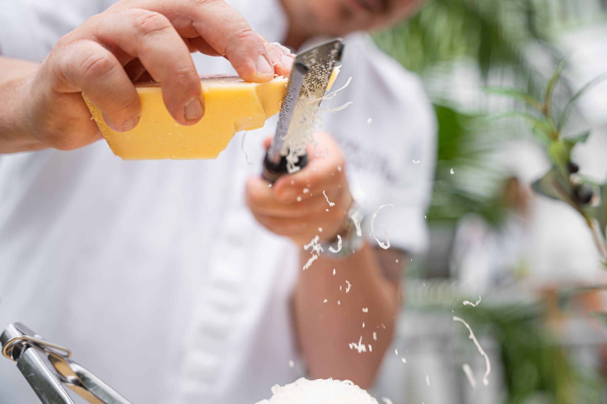 Close-up of hands grating a wedge of parmesan, shavings flying over a dish being prepared outdoors
