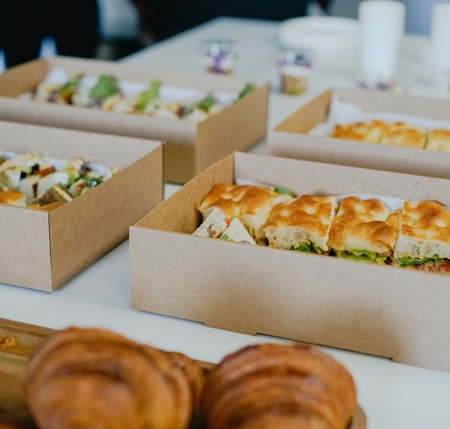 Individual takeaway boxes of stuffed focaccia sandwiches and salad lined up on a table, with bread rolls in the foreground