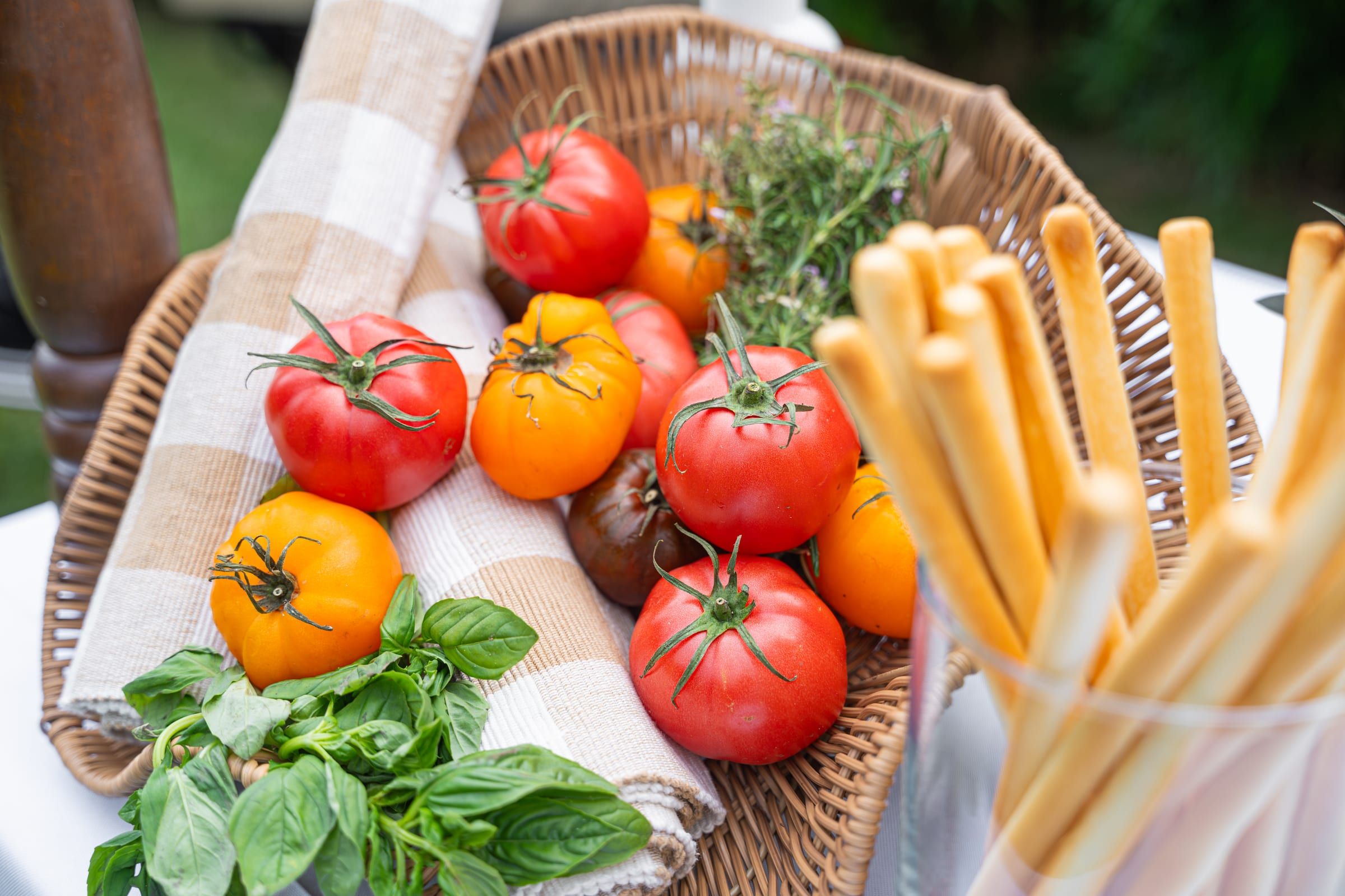 A wicker basket of heirloom tomatoes and fresh basil beside a glass of grissini breadsticks on a checked cloth