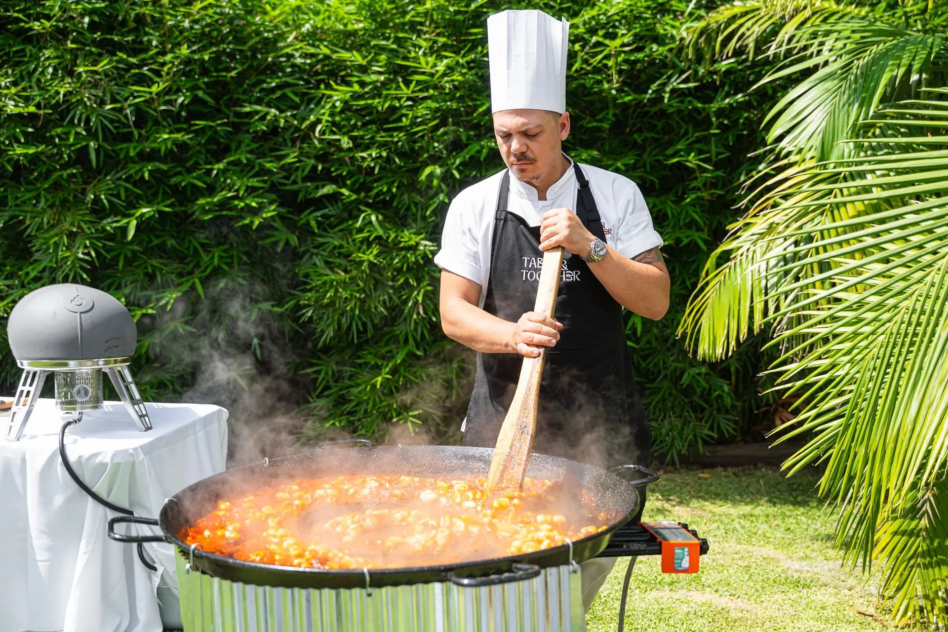 Chef in a Table & Together apron stirring a large pan of tomato sauce and gnocchi at an outdoor live cooking station