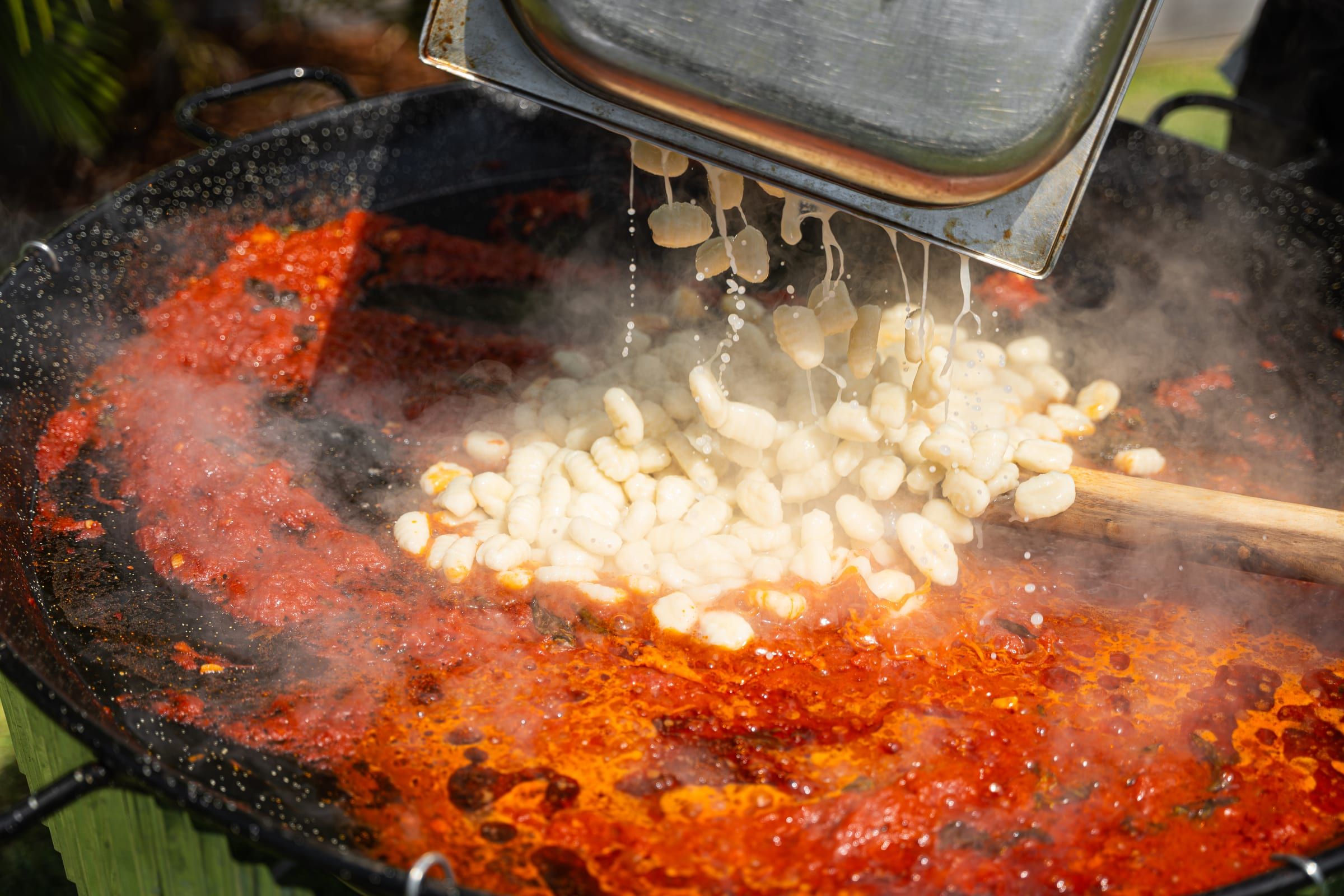 Fresh gnocchi being poured from a tray into a large sizzling pan of tomato sauce on an outdoor live cooking station, steam rising