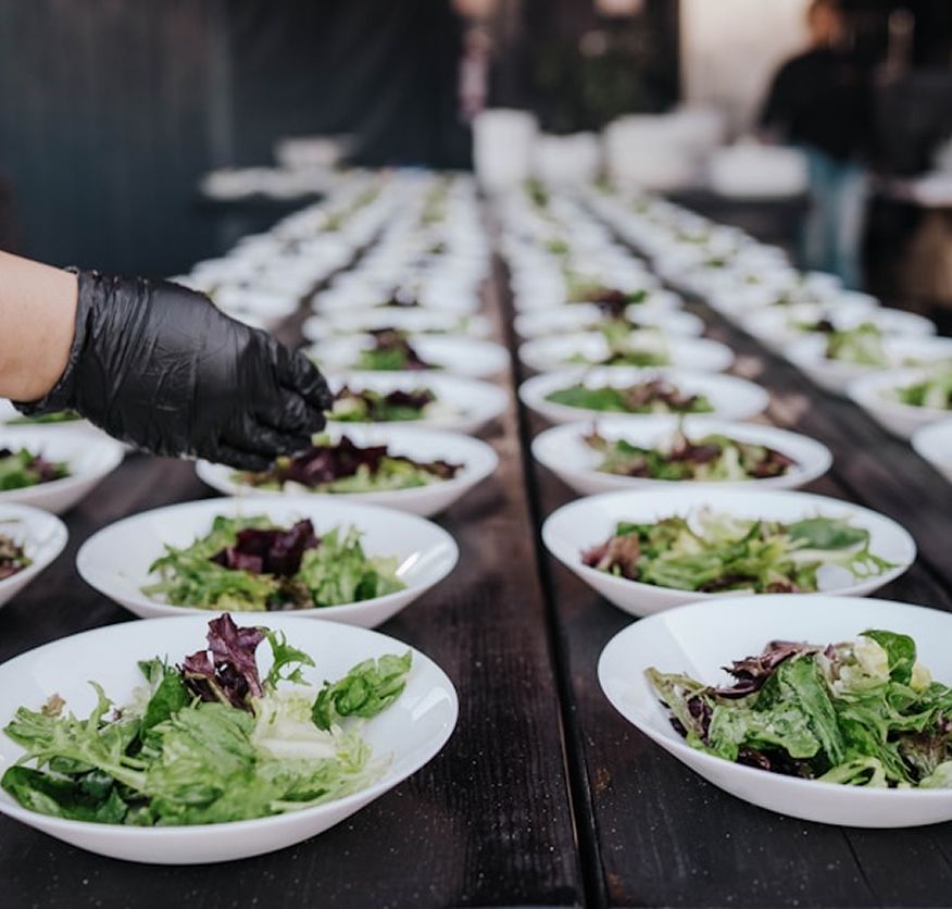 A gloved hand plates mixed leaf salad into a long line of white bowls ahead of service