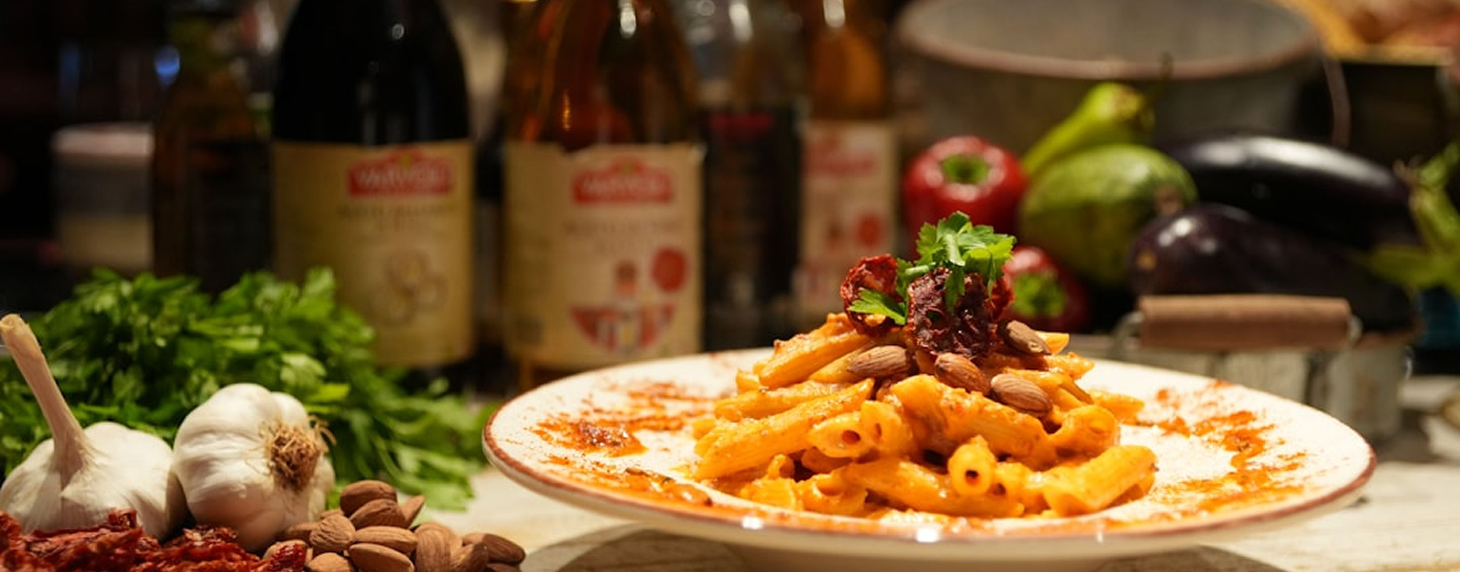A plate of penne in a creamy tomato sauce topped with toasted almonds and sun-dried tomato, styled with wine bottles, garlic bulbs and parsley