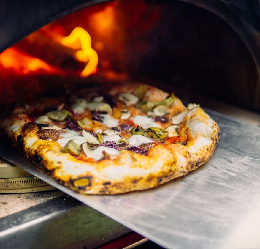 A pizza on a peel in front of the flames of a wood-fired oven