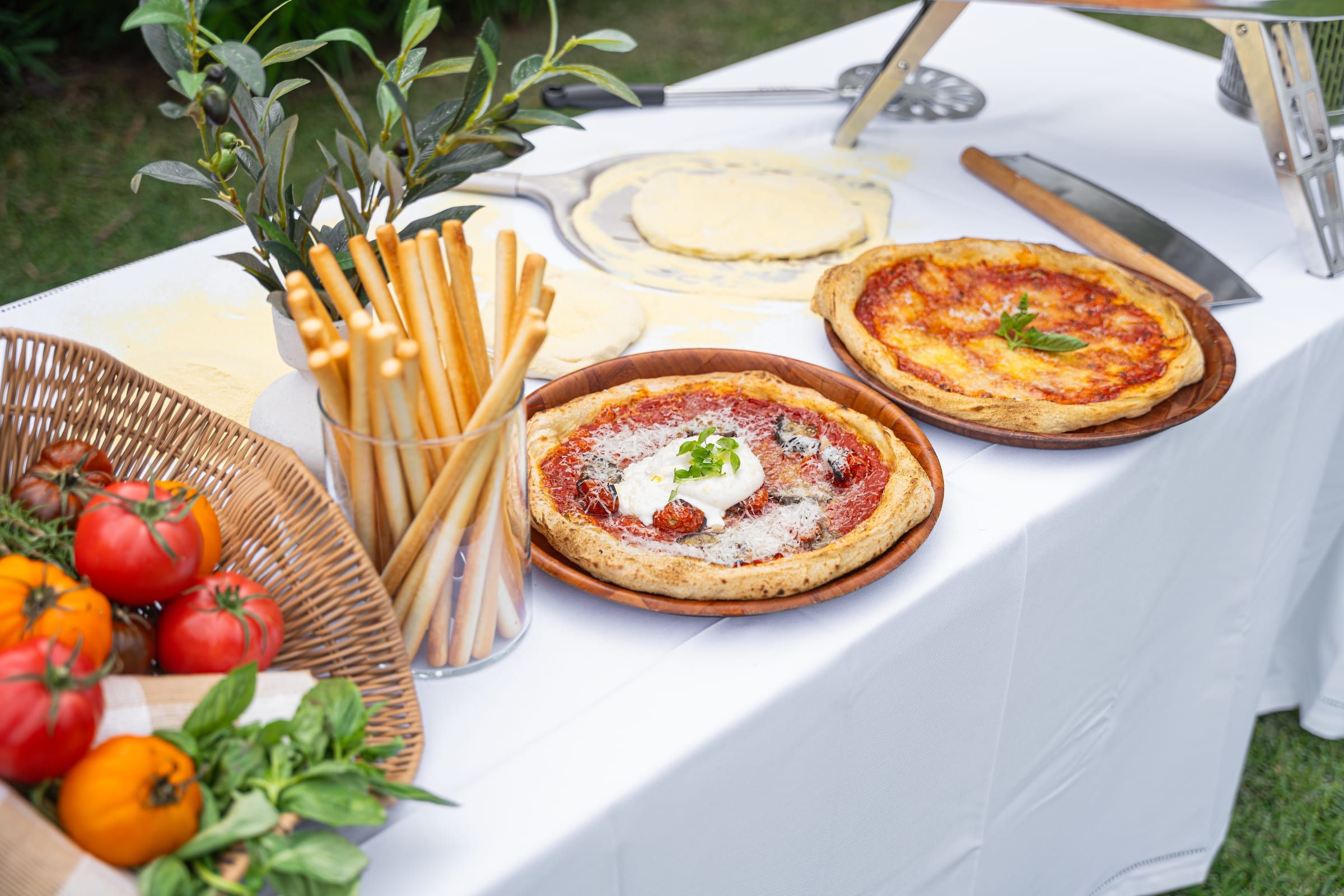 Two burrata pizzas on wooden boards beside a glass of grissini breadsticks and a basket of heirloom tomatoes, with fresh dough being rolled out on the table behind