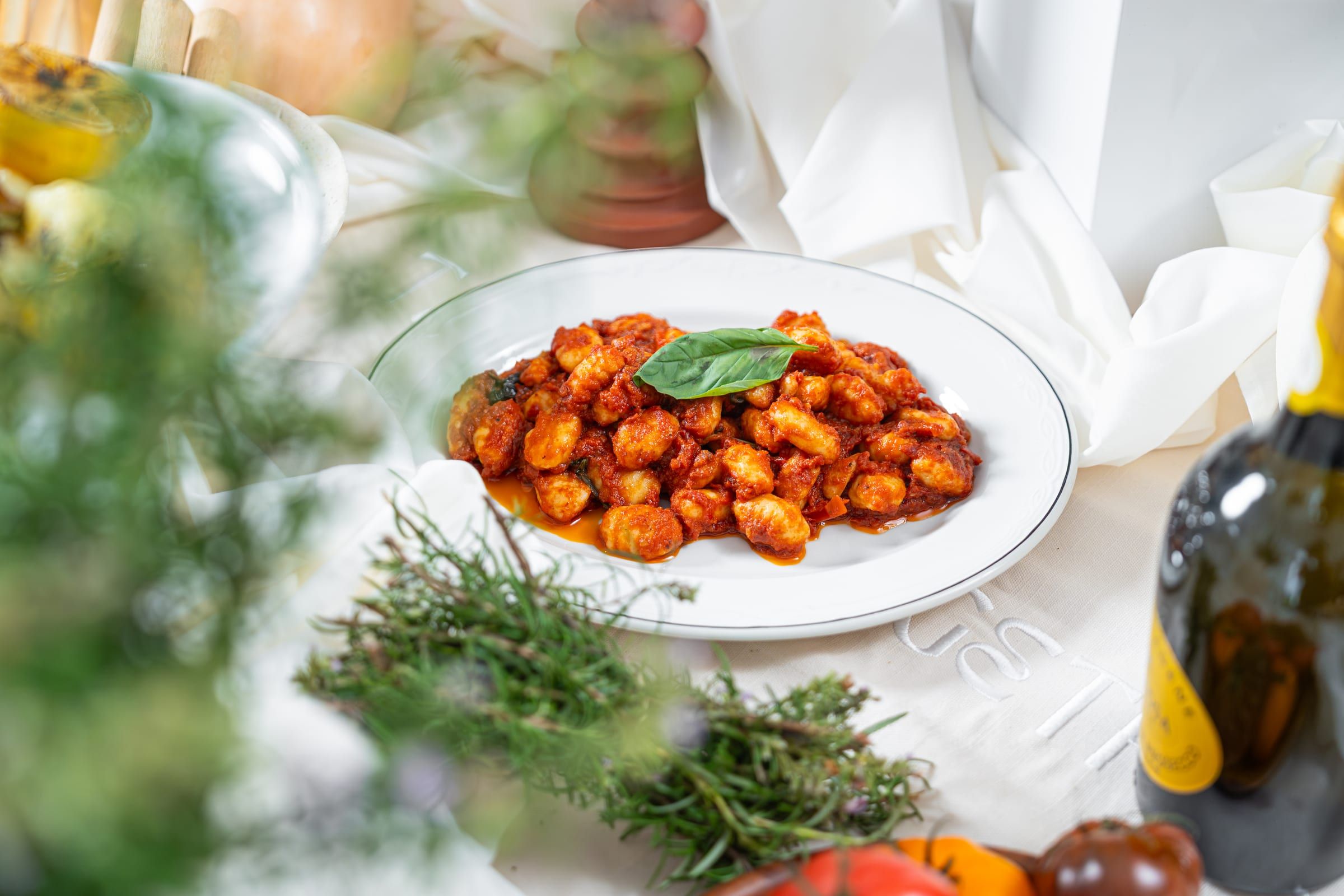A white oval plate of gnocchi in tomato sauce garnished with a basil leaf, set on an embroidered white tablecloth surrounded by fresh herbs, heirloom tomatoes and a bottle of olive oil