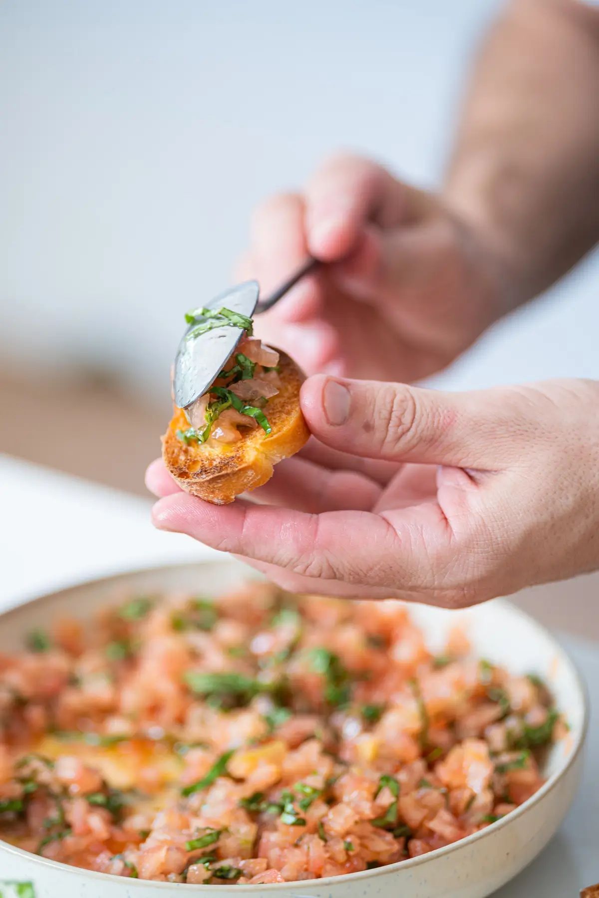 A hand holds a piece of toasted bruschetta while tomato and basil topping is spooned on top, with a bowl of the topping below