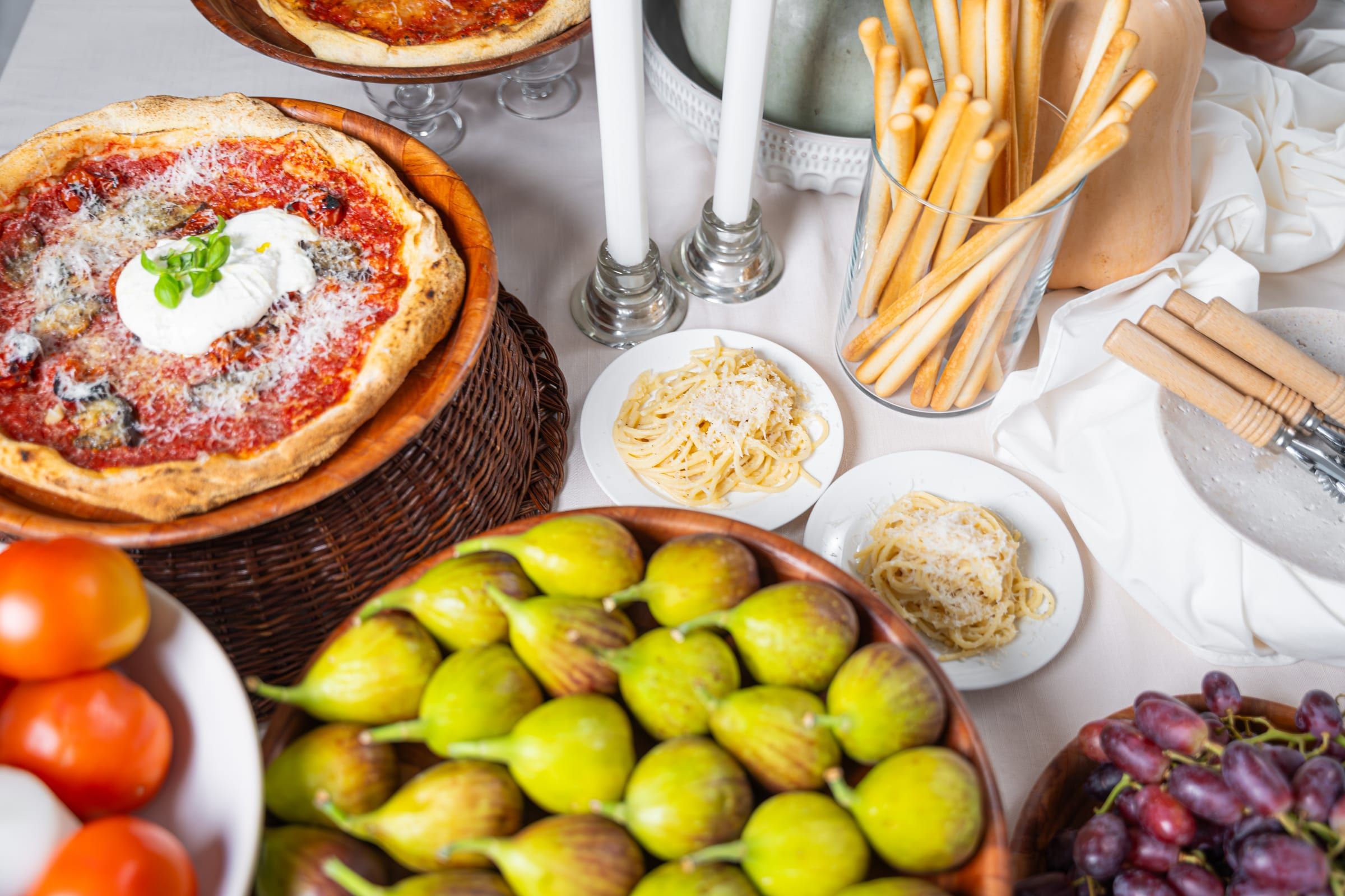 An overhead spread of burrata and tomato pizza, cheese-topped spaghetti, grissini breadsticks and a bowl of fresh figs set out for guests to help themselves