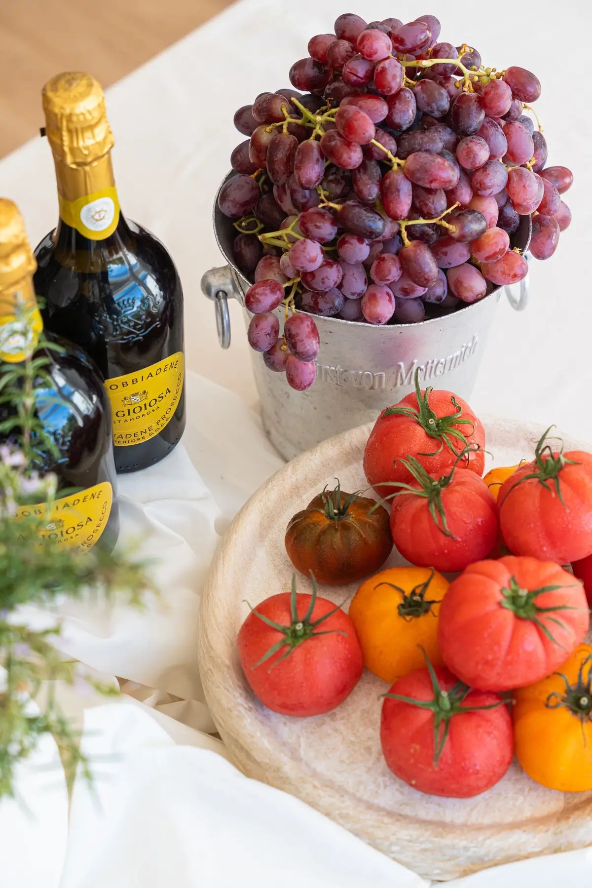 Bottles of Prosecco beside a bucket of grapes and a bowl of heirloom tomatoes, styled on a linen cloth