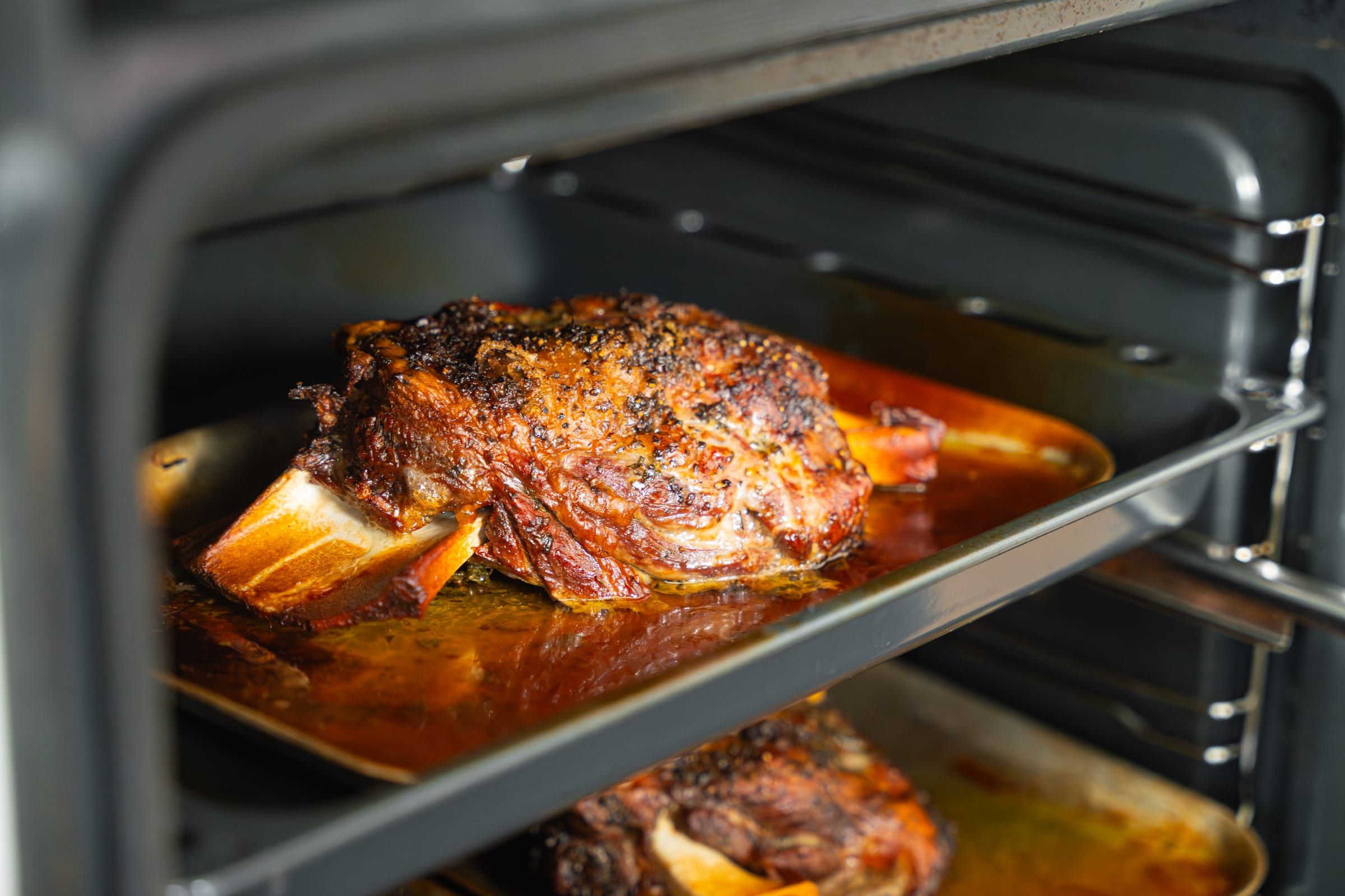 A roasted joint of meat with a deeply charred herb and pepper crust rests on a tray inside a large oven, another tray visible below
