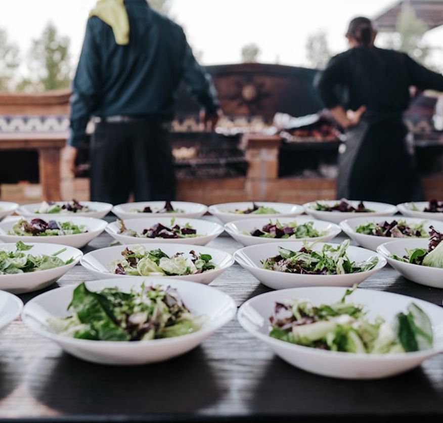 Rows of mixed leaf salad bowls set out on a table in front of an outdoor wood-fired grill, with staff tending the fire behind