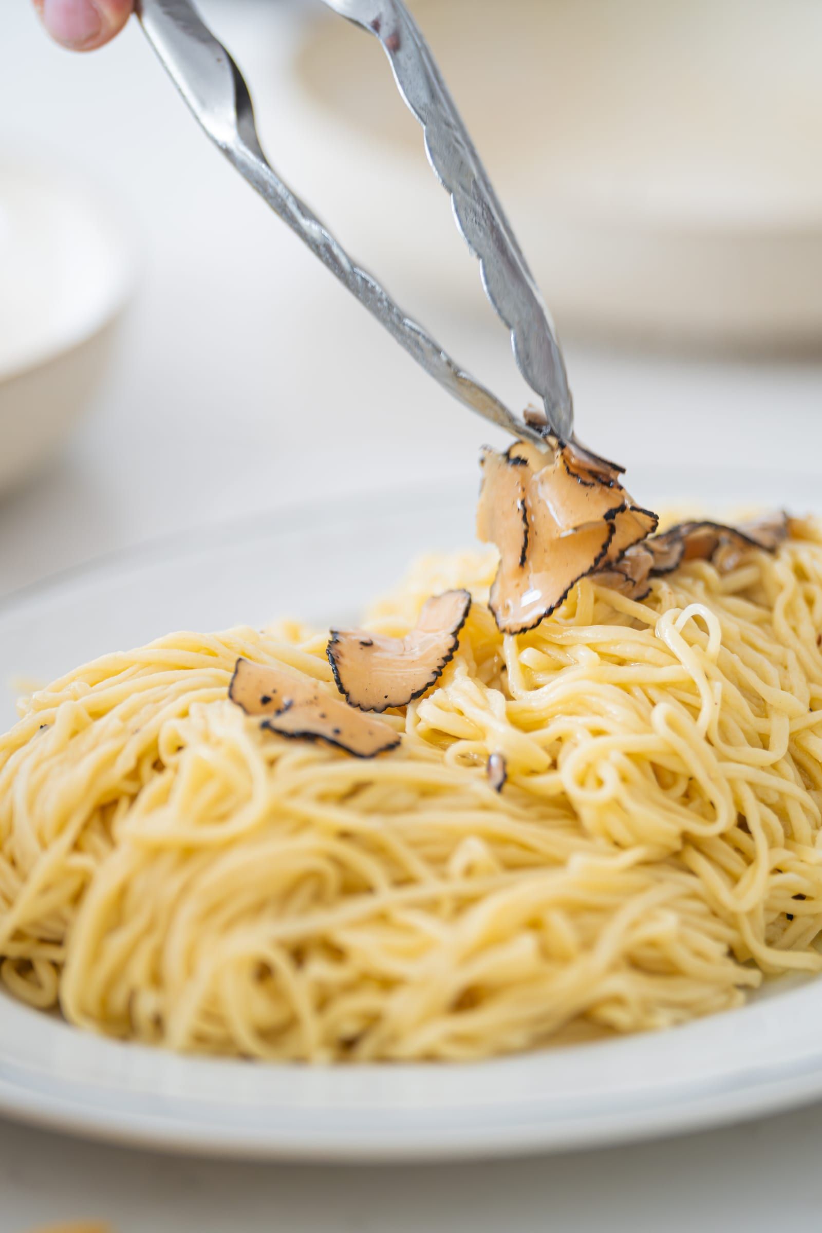 Tongs placing shaved black truffle over a plate of tagliolini pasta
