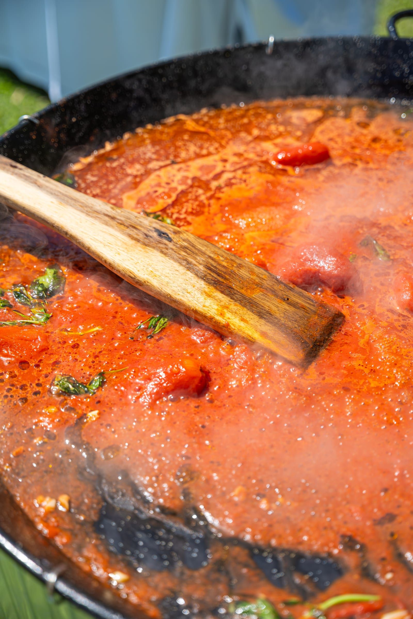 A wooden spoon stirs a large pan of steaming tomato sauce flecked with basil