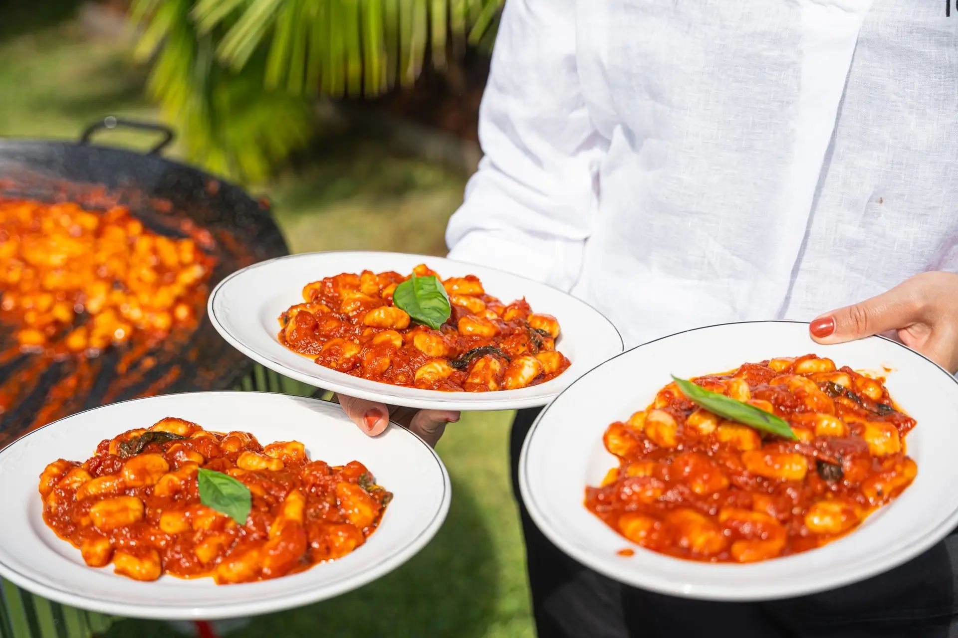 A waiter in white carries three plates of gnocchi in tomato sauce garnished with basil, with the cooking pan visible behind