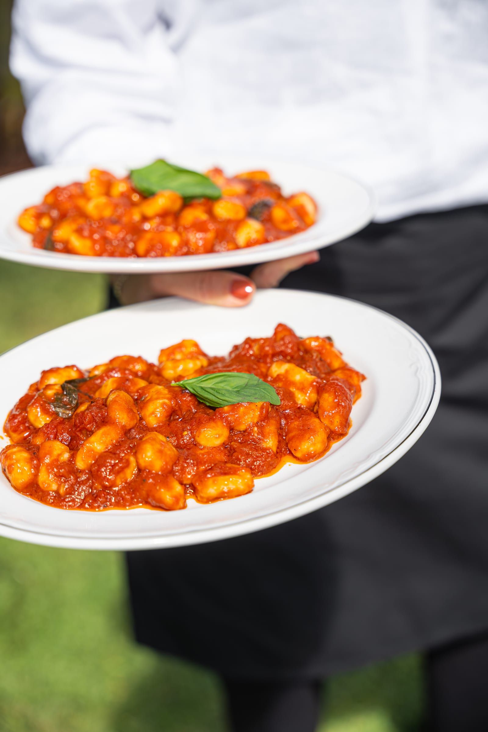 A waiter in a white shirt and black apron holding two plates of gnocchi in tomato sauce garnished with basil, one in each hand, outdoors
