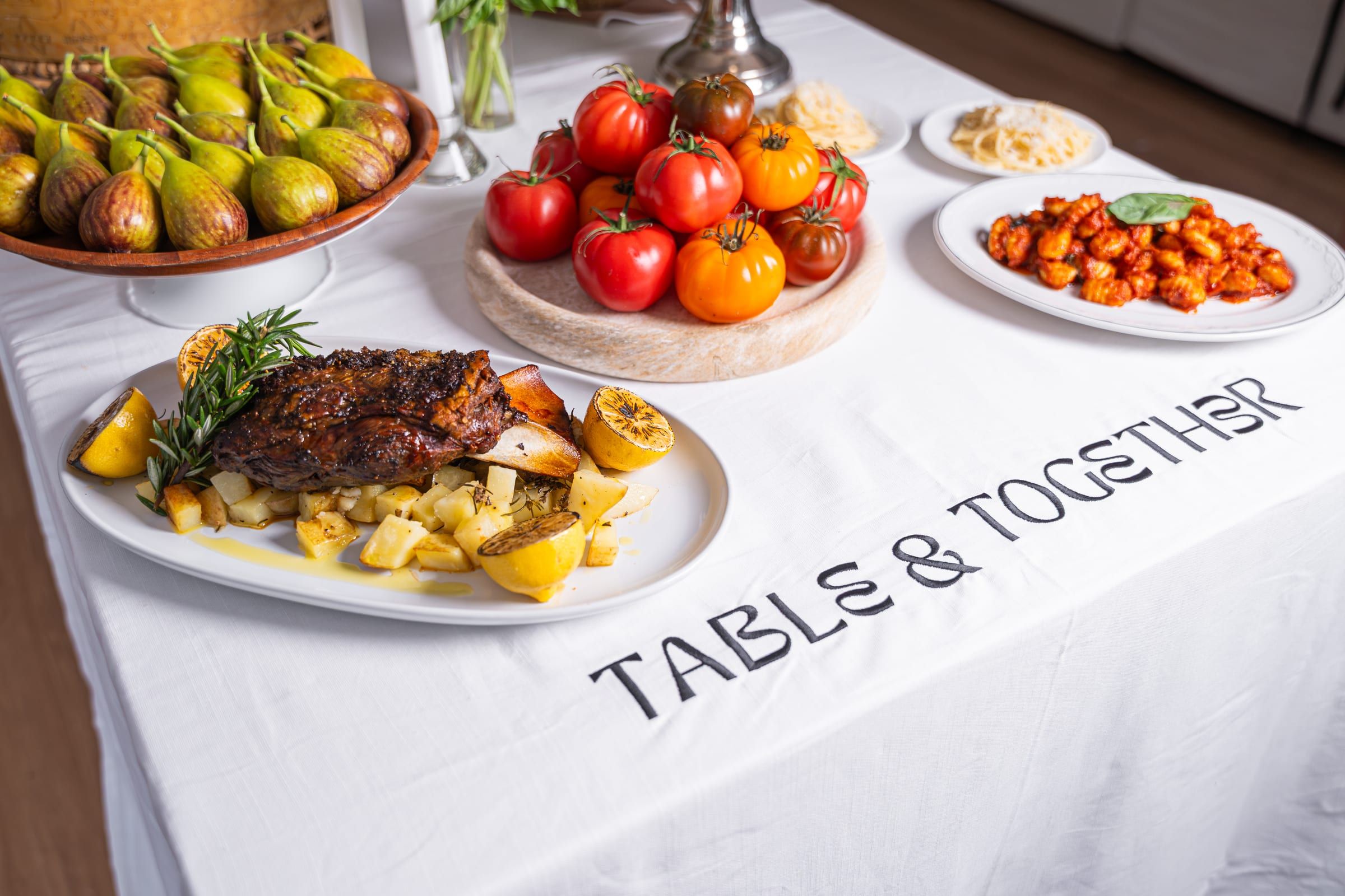A table spread on a Table & Together embroidered tablecloth with a roasted lamb shoulder, charred lemon and rosemary over potatoes, a bowl of figs, a platter of heirloom tomatoes and a plate of gnocchi in tomato sauce